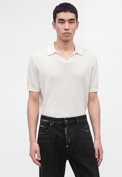 Homme portant une chemise blanche à manches courtes avec col et un jean noir, debout devant un fond clair uni.