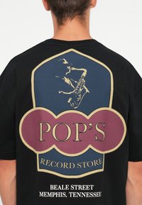 Zwarte t-shirt met een kleurrijke print van een muzikant die saxofoon speelt, met de tekst "POP'S RECORD STORE" en "BEALE STREET, MEMPHIS, TENNESSEE".