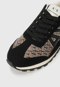 Black sneaker features a textured black suede upper, geometric brown and cream pattern, flat black laces, and a rubber sole.