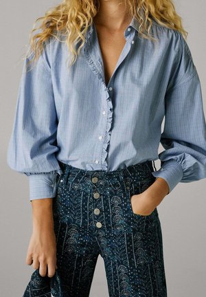 Vrouw draagt een lichtblauw geruite blouse met ruchtdetail en een hooggetailleerde donkerblauwe broek met bloemenpatroon en knopen, één hand in de zak.