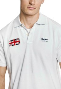 Biela polo košeľa vyrobená z bavlny, so záplatom britskej vlajky na ľavom prsníku a vyšitým nápisom "Pepe Jeans London W11" v námornej modrej.