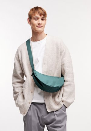 Borsa a tracolla in tessuto teal con forma arrotondata, fissata da una tracolla coordinata, indossata sopra una camicia bianca e un cardigan crema.