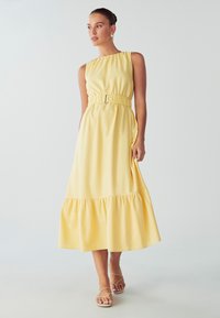 Keltainen maxi-mekko, jossa on muotoiltu vyötärö ja levenevä helma. Yhdessä olkaimessa ja pehmeässä, tekstuurissa kankaassa. Yhdistetty hihnatossujen kanssa.