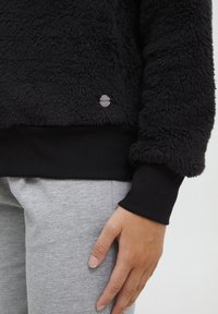 Czarny polarowy golf z teksturowaną powierzchnią i prążkowanymi czarnymi mankietami, z małą srebrną metką logo po lewej stronie blisko dołu.