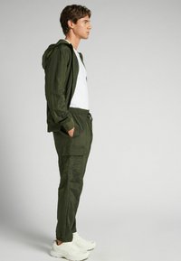 Groene lichtgewicht jack met capuchon, gecombineerd met bijpassende joggers met een elastische tailleband en zijzakken, aangevuld met witte sneakers.