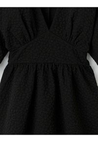 Robe noire présentant un motif texturé matelassé avec un décolleté plongeant en V et des manches froncées, soulignée par des paillettes discrètes sur toute la surface.