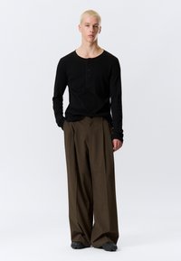 Camisa henley negra de manga larga con tres botones, combinada con pantalones anchos de rayas marrones; modelo de pie con las manos en los bolsillos.