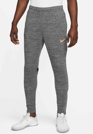 Mann trägt graue Nike-Jogginghosen mit einem pfirsichfarbenen Nike-Logo am Oberschenkel und weißen Sportschuhen, steht vor einem einfarbigen Hintergrund.