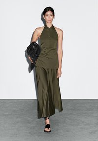 Olivenfarvet, ærmeløs halterkjole med knapper; svajende nederdel. Modellen holder en sort, tekstureret clutch og har sorte sandaler på.