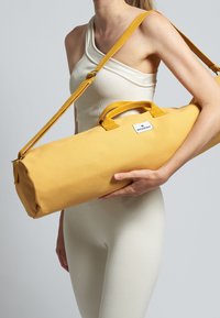 Bolsa de deporte de lona amarilla con dos asas y una correa para el hombro. Presenta una etiqueta de logo blanca en la parte frontal y tiene forma cilíndrica.