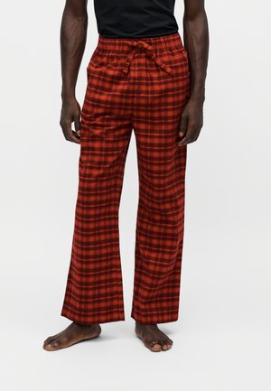 Homme portant un pantalon ample à carreaux rouges et noirs avec une taille à cordon, debout pieds nus devant un fond uni.