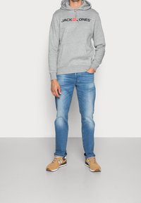 Felpa grigia in cotone con logo frontale, jeans blu e sneakers beige con suola bianca e accenti scuri. Outfit informale per l'uso all'aperto.