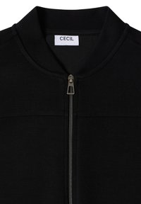 Veste noire à fermeture éclair avec col montant et tirette en argent, avec une étiquette de marque blanche "CECIL" à l'intérieur du col.