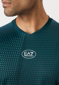 Türkiz színű sportpóló V-nyakkal, áttűnő pöttyös mintával és EA7 Emporio Armani logóval a bal mellrészen. Sima textúra.