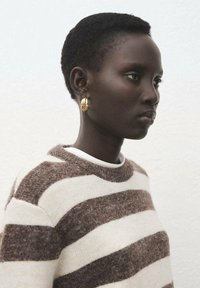 Perfil de una persona con cabello rizado corto que lleva pendientes de aro dorados y un suéter peludo a rayas marrón y blanco, sobre un fondo liso.