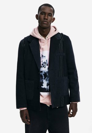 Schwarze Jeansjacke mit kontrastierenden Nadelstreifen, zwei Fronttaschen, darüber eine rosa Kapuzenjacke mit grafischem Druck und Rundhalsausschnitt.