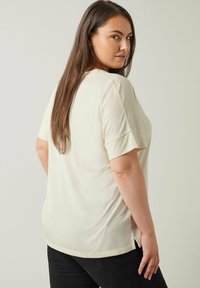 Lyhyt hiha vaalea beige paita, jossa on rento istuvuus. Paidassa on pyöreä kaula-aukko ja sivuhalkiot. Kangas näyttää pehmeältä ja sileältä.