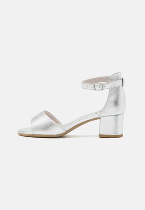 Stříbrná sandál s otevřenou špičkou a přezkou na zápěstí s nízkým blokovým podpatkem na bílém pozadí.