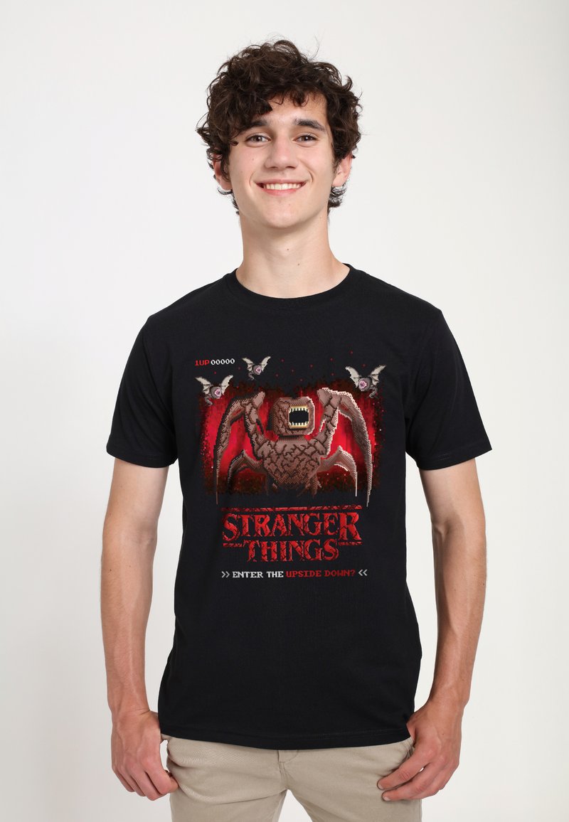 Camiseta negra de algodón con un gráfico de un monstruo y murciélagos en tonos rojos, con el texto "STRANGER THINGS" y elementos de diseño inspirados en videojuegos.