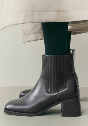 Bottines en cuir noir à talons blocs portées avec des collants foncés et l'ourlet d'un manteau beige visible au-dessus.