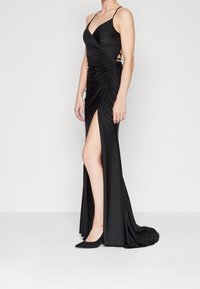 Černé večerní šaty s přiléhavou siluetou, nařaseným designem, hlubokým výstřihem a rozparkem až ke stehnům, v kombinaci s černými lodičkami se špičatou špičkou.