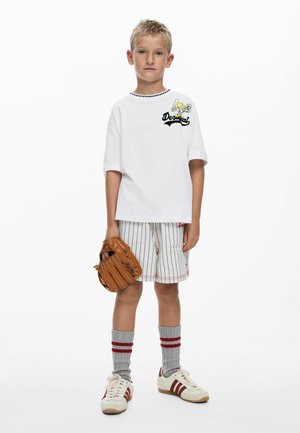 Witte T-shirt met korte mouwen en zwart "Desigual"-logo, gestreepte korte broek, grijze sokken met rode strepen, en witte schoenen met rode strepen, die een bruine handschoen vasthoudt.