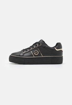 Black low-top sneaker with thick sole, gold eyelets, side logo, and gold accent on heel, photographed on white background.