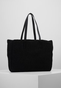 Svart tygväska tillverkad av texturerad fleece med två svarta läderhandtag. Har en rektangulär form med en mjuk, plyschyta.