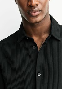 Melnā ribota krekliņa ar struktūrētu apkakli, divām pogām un teksturētu audumu ar vertikālām līnijām. Tuvplāns apkakles zonai.