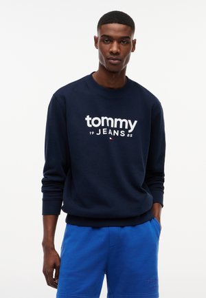 Młody mężczyzna w granatowej bluzie z kapturem z logo "tommy JEANS" w białym kolorze oraz niebieskich szortach, stojący z jedną ręką w kieszeni na jednolitym tle.