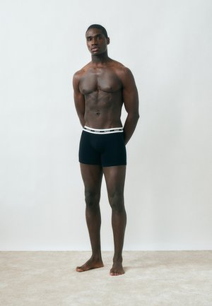 Muskuløs mann stående barbeint på teppe, iført svarte boxer briefs med hvit midjebånd merket "HUGO", hendene bak ryggen, nøytral bakgrunn.