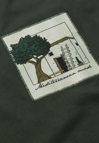 Groene katoen stof met een geborduurd label dat een boom, een wit gebouw en een ladder afbeeldt, met de tekst "Mediterranean mode."