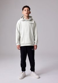 Sudadera con capucha gris claro con bolsillo delantero y logo negro, combinada con pantalones jogger negros con un logo. Zapatillas blancas completan el conjunto.