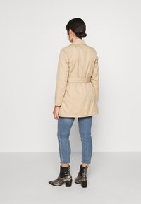 Trench-coat beige avec une taille ceinturée, des manches longues et un col à revers, associé à un jean skinny bleu et des bottines à motif.