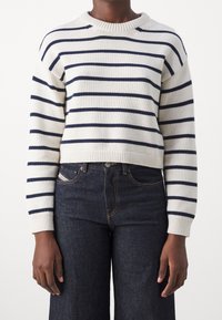 Maglione bianco a maglia con righe orizzontali blu navy, polsini a costine e scollo rotondo. Abbinato a jeans in denim scuro.