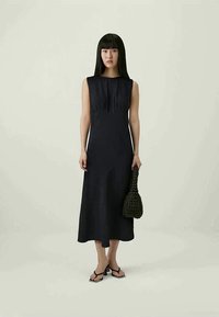 Crna haljina bez rukava s prilagođenim korzetom i lepršavom suknjom, s nabranim detaljima. Manekenka drži teksturiranu, tamnu torbu od tkanine i nosi jednostavne sandale.