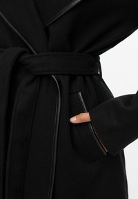 Czarny płaszcz z skórzanymi detalami i paskiem zawiązanym w talii, ręka częściowo włożona w boczną kieszeń na tle jednolitego jasnego koloru.