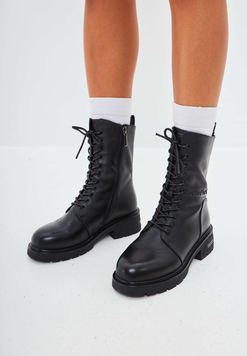 Botas de combate de cuero negro con punta redonda, cierre de cordones en la parte delantera, cremallera lateral y suela gruesa. Presentan costuras y un detalle del logotipo en el lateral.