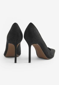 Scarpe con tacco alto in suede nero, con punta affusolata, sottile tacco a stiletto e dettagli di chiusura aperta nella parte frontale, che mettono in evidenza una texture liscia.