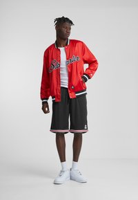 Chaqueta de satén rojo tipo varsity con acentos negros y logo "Starter", combinada con pantalones cortos negros con rayas rojas y zapatillas blancas.