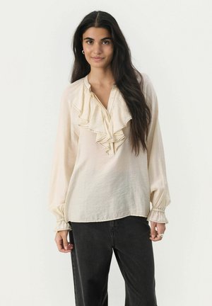 Krämfärgad blus med volang fram, långa ärmar med elastiska manschetter och knappstängning i halsen. Kombinationen med mörka jeans som har en subtil textur.