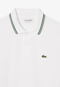 Polo blanc avec col rayé vert, deux boutons, et logo crocodile vert brodé sur la poitrine gauche. Design à coupe régulière.