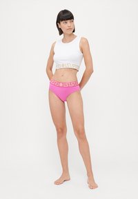 Balts cropped tops ar zeltītu grieķu atslēgu rakstu; rozā augstās vidukļa bikses ar atbilstošu jostasvietu. Gluda auduma, minimālas šuves, pieguļošs dizains.
