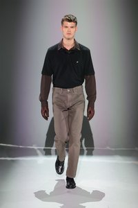 Zwarte poloshirt met donkerbruine mouwen, beige broek en zwarte schoenen. Het model loopt op een effen, goed verlichte achtergrond.