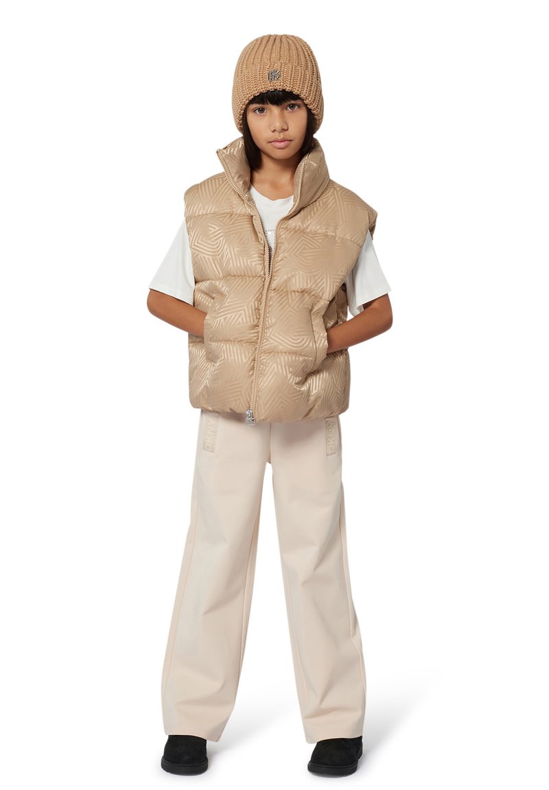 Beige gewatteerde puffer vest met een geometrisch patroon, gecombineerd met een wit T-shirt, beige wijde broek en een bruine gebreide beanie.