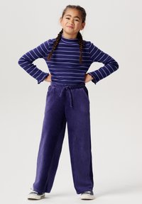 Lila randig långärmad topp med volangärmar, kombinerad med vida kordbyxor i enhetlig lila. Vita sneakers fulländar stilen.