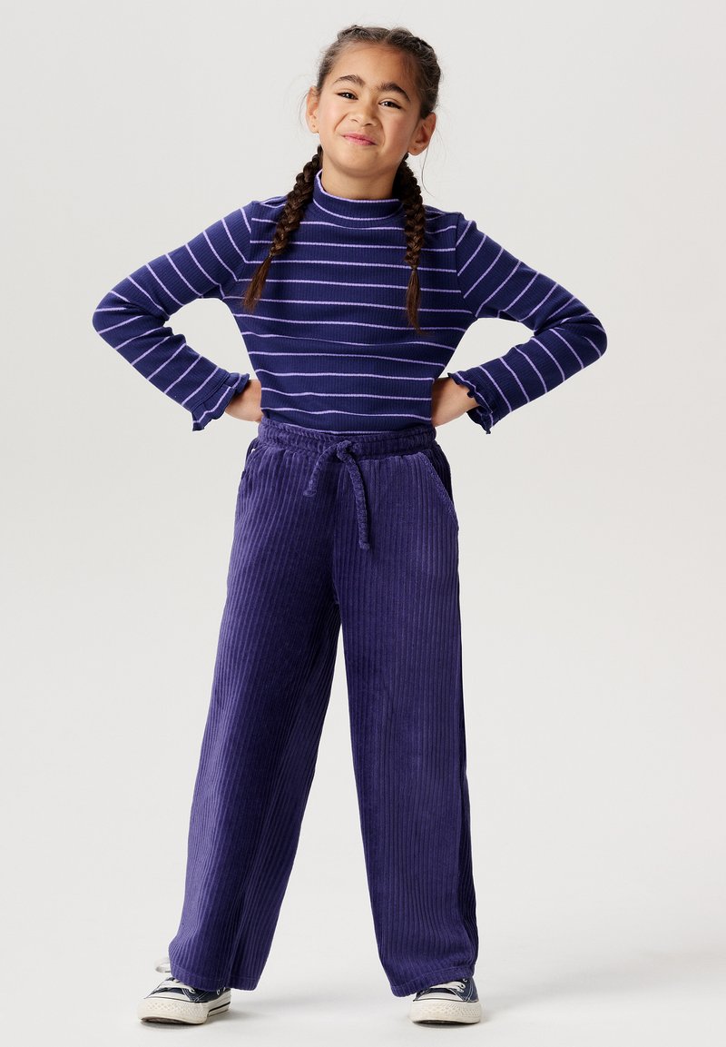 Lila randig långärmad topp med volangärmar, kombinerad med vida kordbyxor i enhetlig lila. Vita sneakers fulländar stilen.