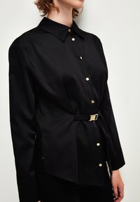 Maglietta nera a maniche lunghe realizzata in tessuto liscio, con colletto, bottoni dorati e un design con cintura laterale e orlo diagonale.