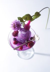 Fioletowa szklana butelka z różową pipetą, oznaczona etykietą "Equal Berry Bakuchiol Plumping Serum", otoczona fioletowymi jagodami i srebrną łyżeczką.
