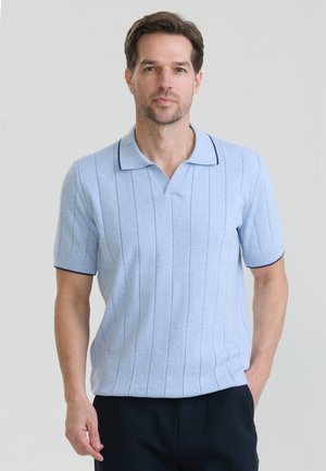 Mężczyzna o jasnobrązowych włosach, ubrany w jasnoniebieską koszulkę polo z krótkim rękawem i cienkimi pionowymi paskami oraz ciemne spodnie, stojący na jednolitym tle.